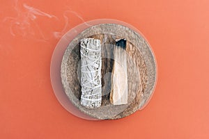 Burning incense bundle - white sage and palo santo stick with smoke on wooden stand over orange brown background