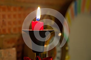 Burning red candle in black candelabra. Stands on brick fireplace. Clocks on background