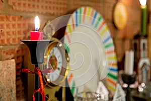 Burning red candle in black candelabrum tied with red ribbon. Stands on brick fireplace. Old retro clocks and white plate on