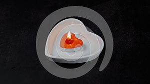 Burning red candle heart in a white saucer on a black background