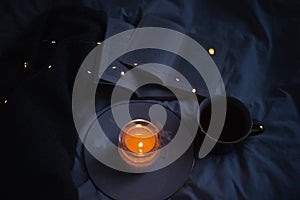 Burning scented candle on ceramic tray with open paper book and knit sweater cloth in bed.