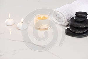 Burning white candles in glass near spa stones and rolled towel on marble white surface