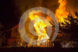 Burning wooden house at night. Bright orange flames and dense smoke from under the tiled roof on dark sky, trees silhouettes and r