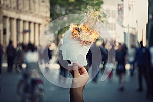 Burnout and stress concept as person hand holds a burning, human head shaped, paper sheet on a crowded street. Memory loss and