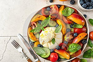Burrata cheese salad with grilled peaches, tomatoes, blueberries, cucumber and basil in a plate with cutlery