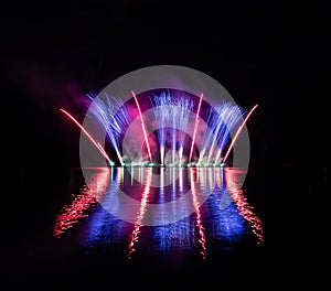 Bursts of blue and red fire in rich fireworks over Brno`s Dam with lake reflection