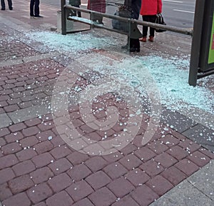 A bus stop with glass broken into small crumbs, shards of glass lie on the pavement, only people`s legs are visible.