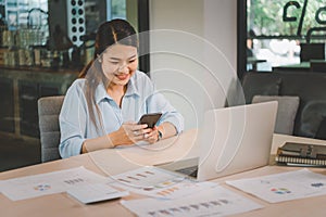 Business asian woman sitting at her desk with laptop using mobile phone during checking an email or social media on internet