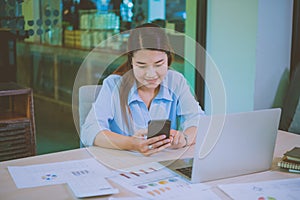 Business asian woman sitting at her desk with laptop using mobile phone during checking an email or social media on internet