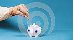 Business banner. Wrinkled hand of an elderly woman puts a euro coin in a piggy bank. Blue background. Copy space. The