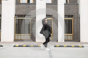 Business beautiful young woman in fashionable black clothes with a formal suit and white shoes is walking near a modern building