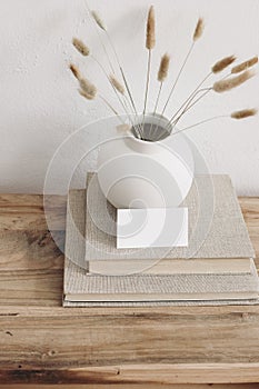 Business card mockup on vintage wooden table. Modern ceramic vase with dry Lagurus ovatus grass and pile of books. White