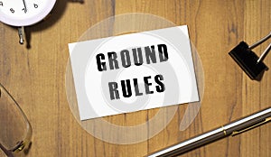 A business card with the text GROUND RULES lies on a wooden office table among office supplies