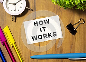 A business card with the text HOW IT WORKS lies on a wooden office table among office supplies. Business concept