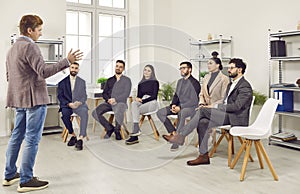 Business coach talking to a team of young people sitting on chairs in the company office