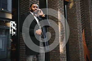 Business communications. Happy young African man in formal wear talking on the mobile phone and smiling while standing outdoors