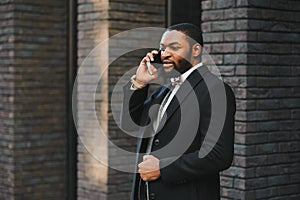 Business communications. Happy young African man in formal wear talking on the mobile phone and smiling while standing outdoors