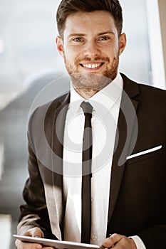 Business concept. Happy smiling young businessman standing in office holding wireless touchpad tablet computer. Man in