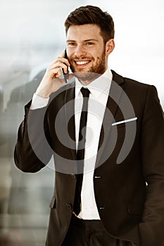 Business concept. Happy smiling young businessman standing in office talking on a cell phone getting good news about his