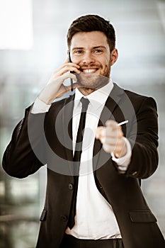 Business concept. Happy smiling young businessman standing in office talking on a cell phone getting good news about his