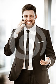 Business concept. Happy smiling young businessman standing in office talking on a cell phone getting good news about his