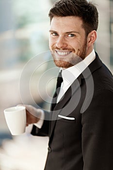 Business concept. Successful young businessman at work. Manager standing in office happy drinking coffee from cup. Man