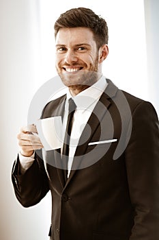 Business concept. Successful young businessman at work. Manager standing in office happy drinking coffee from cup. Man