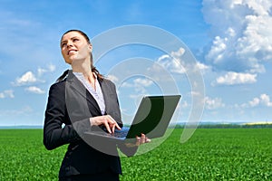 Business concept - woman in green grass field outdoor work on laptop. Young girl dressed in black suit. Beautiful spring landscape