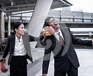 Business concept : Young Asian business woman in suit uniform wearing boxing gloves and punching business man`s face.