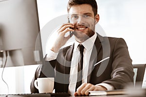 Business concept. Young businessman sitting at the office table happy talking on a cell phone getting good news about