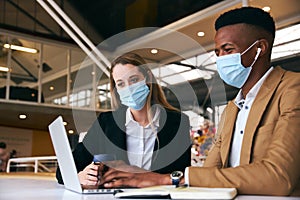 Business Couple Wearing Masks Using Laptop At Hot Desk In Modern Office During Health Pandemic