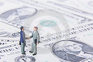 Business deal or agreement and success concept. Two miniature businessmen shaking hands while standing on american dollar money ba
