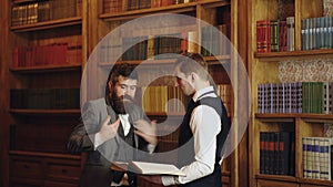 Business employees chatting in library. Two men reading book in the hand on bookshelf background. Men conversation. Law