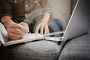 business executive working on laptop and writing on a notebook while sitting sofa at home office