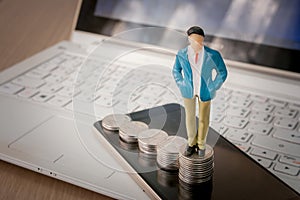 Business man standing on top of stack of silver coins and smartphone with laptop in the background.