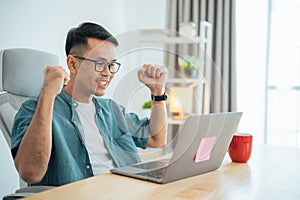 Business freelance asian man wearing glasses smile and happy with success while working online on laptop and celebrating at home