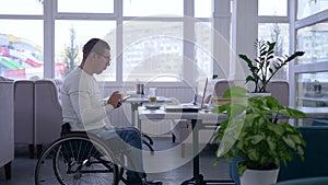 Business freelance, senior man crippled in wheelchair wearing glasses uses a mobile phone and working on laptop sitting