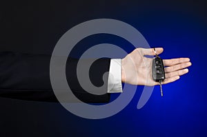 Business and gift theme: car salesman in a black suit holds the keys to a new car on a dark blue background in studio