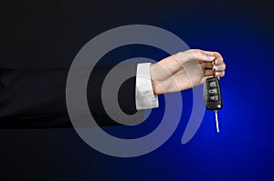 Business and gift theme: car salesman in a black suit holds the keys to a new car on a dark blue background in studio