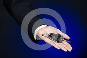 Business and gift theme: car salesman in a black suit holds the keys to a new car on a dark blue background in studio