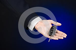 Business and gift theme: car salesman in a black suit holds the keys to a new car on a dark blue background in studio