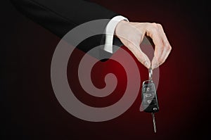 Business and gift theme: car salesman in a black suit holds the keys to a new car on a dark red background in studio