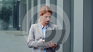 Business on go. Confident businesswoman typing on laptop, standing outdoors near office building, free space