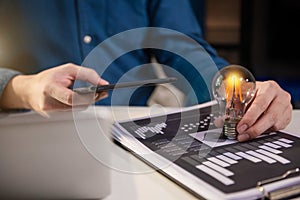 Business hand holding lightbulb with using smartphone, laptop computer and money stack in office.