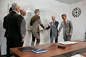 Business handshake. Two happy businessmen in classic wear shaking hands while having a meeting with their team in the