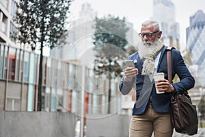 Business hipster senior man using mobile phone while walking to work with city in background - Focus face