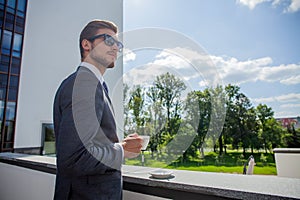 Business, hot drinks and people and concept - young serious businessman with paper coffee cup over office building.