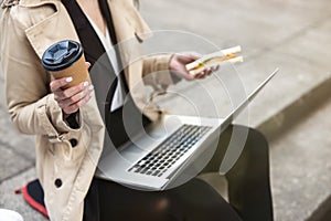 Business lady holding laptop on her knees with coffee and sandwich in hands working during lunch break on the stairs near office,