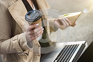 Business lady holding laptop on her knees with cup of coffee and sandwich in her hands sitting near office during lunchbreak ,