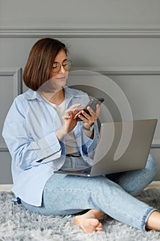 A business lady with glasses on her eyes, wearing a white t-shirt, blue shirt and light jeans is sitting on a gray carpet on the